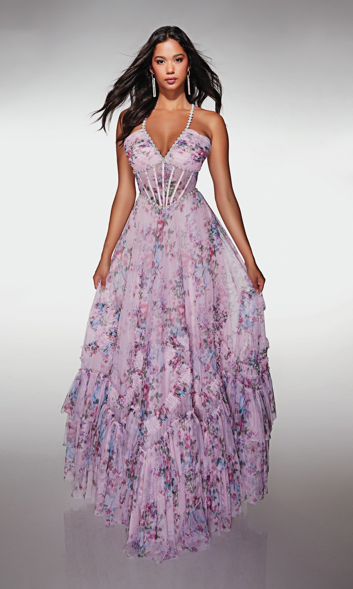 Woman wearing the Alyce 62028 long prom dress in lavender floral print, featuring a sheer corset bodice, thin straps, and a flowing layered A-line skirt, stands against a plain background.